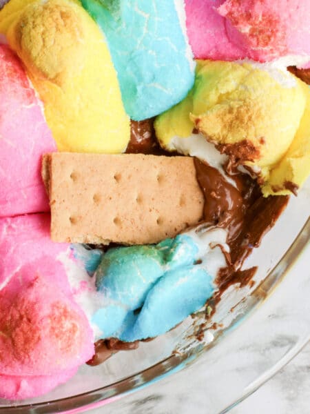A close-up of colorful peeps marshmallows and chocolate melted in a glass dish with a graham cracker dipping into the Easter Peeps smore's dip.