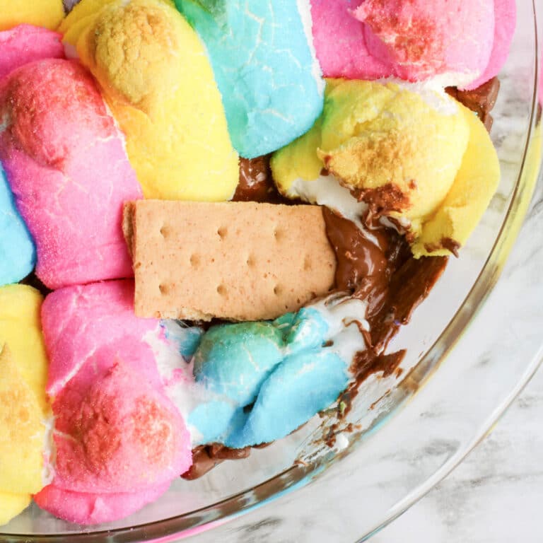 A close-up of colorful peeps marshmallows and chocolate melted in a glass dish with a graham cracker dipping into the Easter Peeps smore's dip.