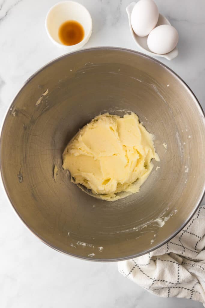 A metal mixing bowl with creamed butter and sugar, next to a small bowl of vanilla extract, and tow eggs for fruit pizza cookies.