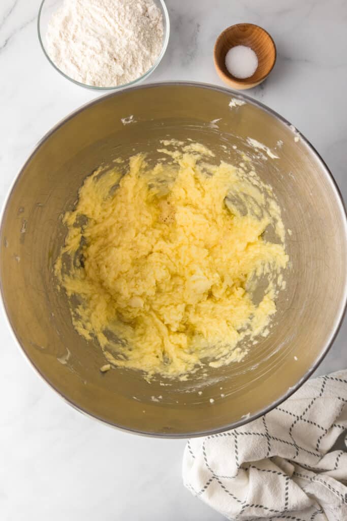 A metal mixing bowl with creamed butter and sugar mixed with vanilla and egg with bowls of flour and salt nearby.