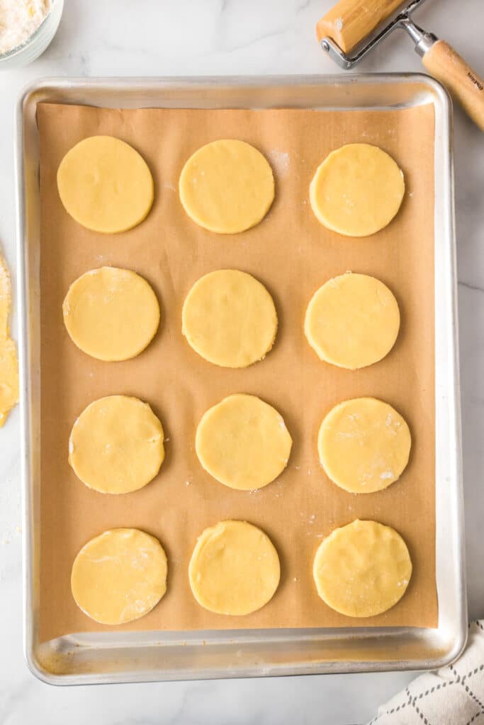 A baking sheet lined with parchment paper holding 12 raw sugar cookies for fruit pizza.