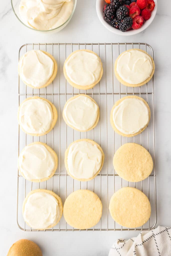 A cooling rack with twelve sugar cookies, eight frosted and four plain with a bowl of fruit nearby for fruit pizza cookies.