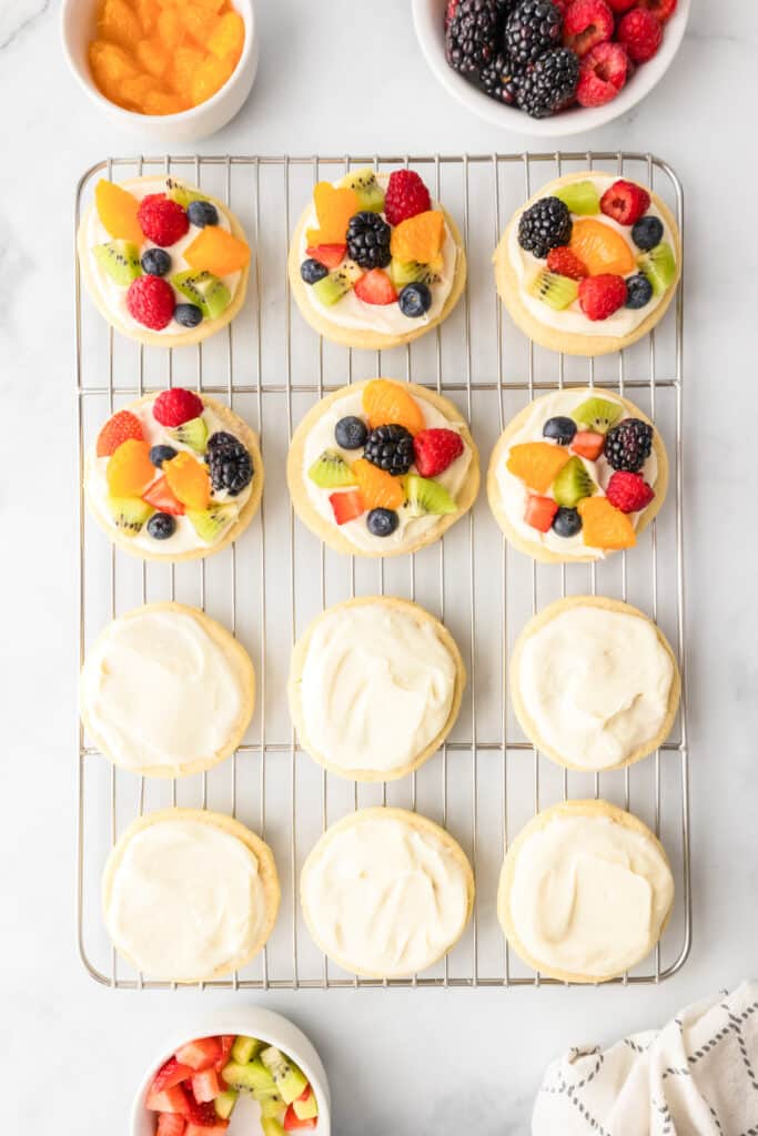 A cooling rack with fruit pizza sugar cookies, some topped with fruit and some with white frosting, surrounded by bowls of fruit on a marble counter.