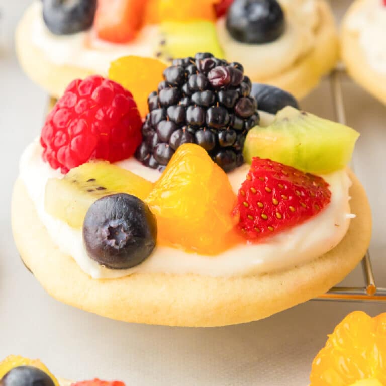 Square view of a fruit pizza suage cookie frosted and topped with fresh fruit from the side on a wire rack.