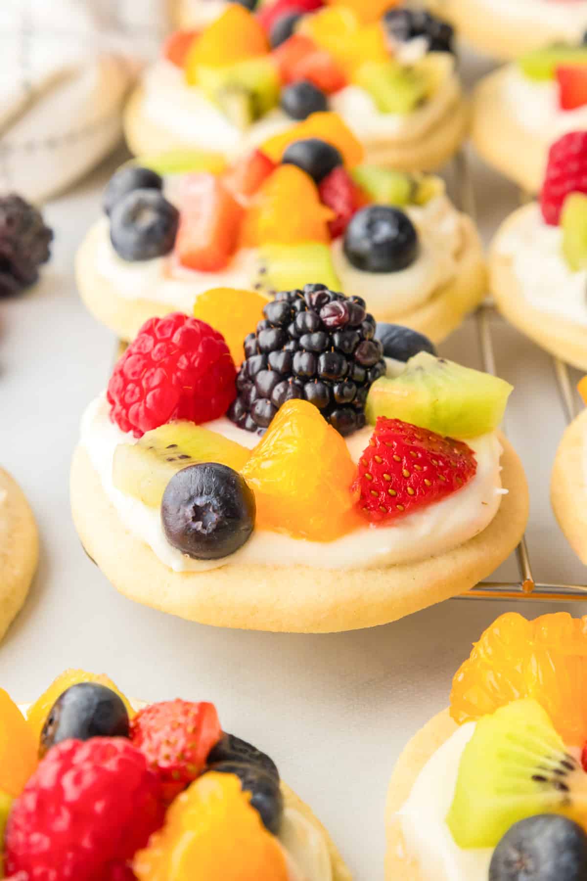Fruit pizza cookie frosted and topped with diced frest fruit from the side on a wire rack with more cookies in the background.