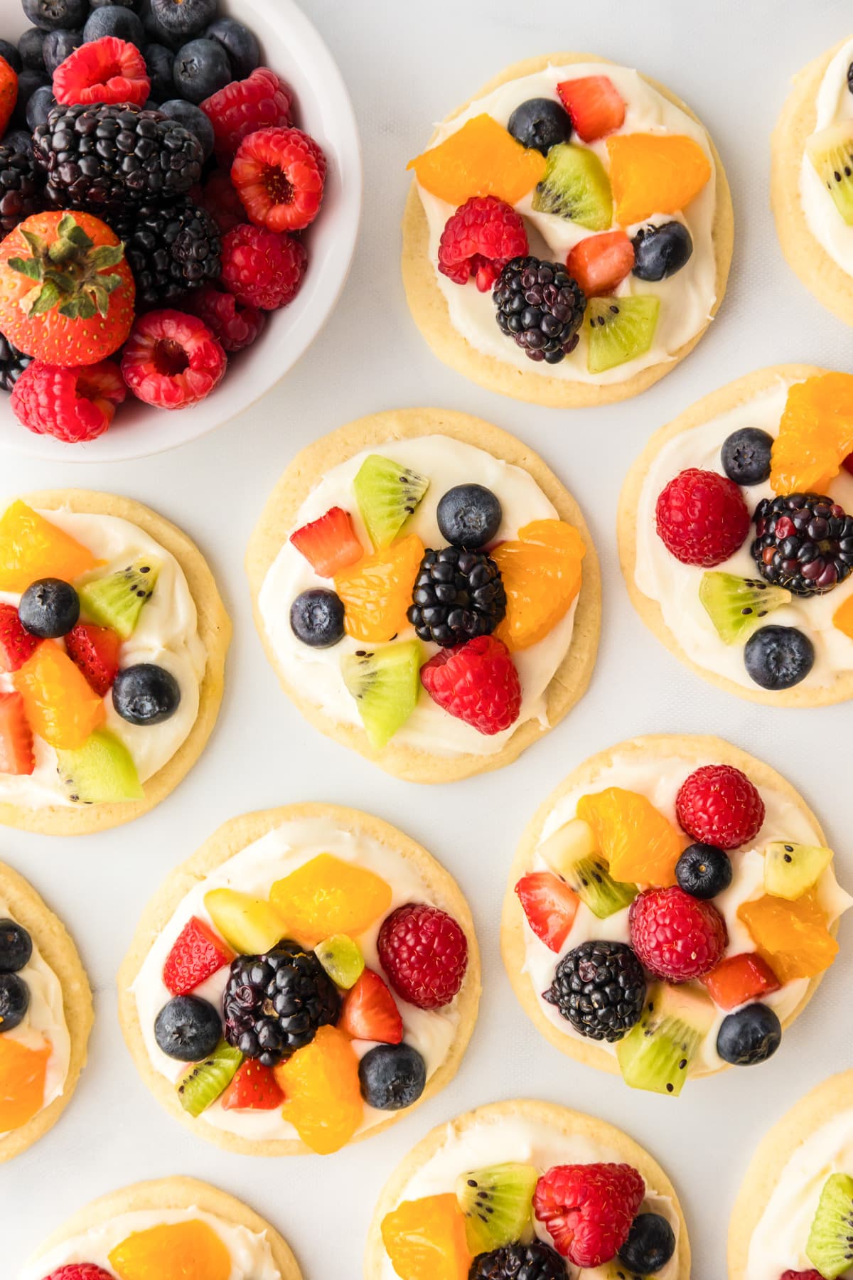 Round fruit pizza sugar cookies topped with frosting and fruit from overhead on a counter after decorating with a bowl of fresh fruit nearby.