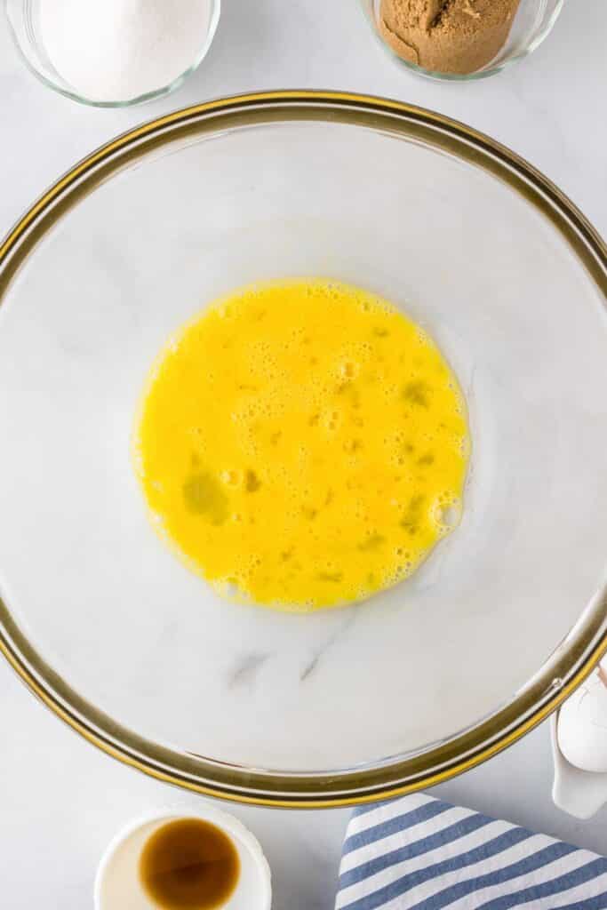 A clear glass bowl with beaten eggs sits on a white surface, surrounded by wet ingredients for brownies.