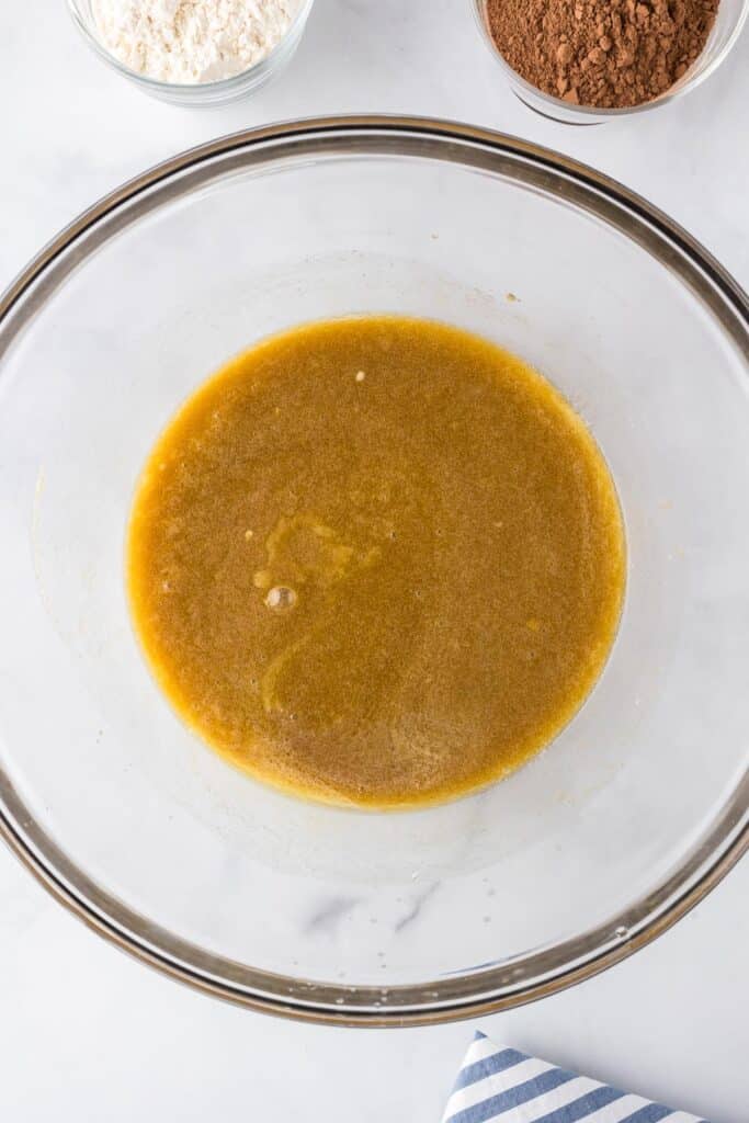 A glass mixing bowl contains all the wet ingredients for peanut butter brownies with dry ingredients nearby on the counter.