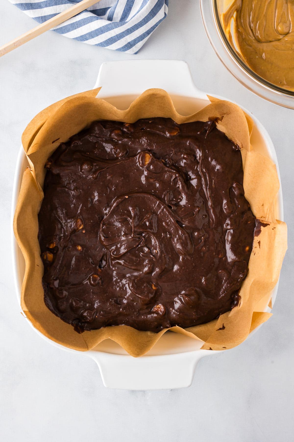 A square baking dish lined with parchment paper filled with brownie batter with peanut butter in a bowl nearby.