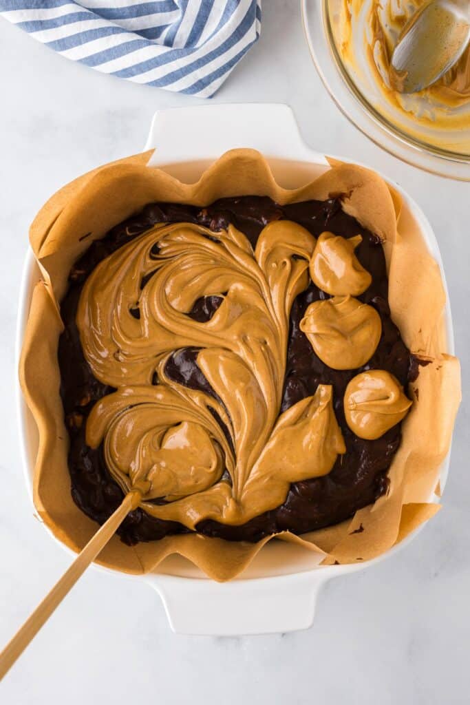 A square baking dish lined with parchment paper full of brownie batter with a chopstick swirling peanut butter through the batter.