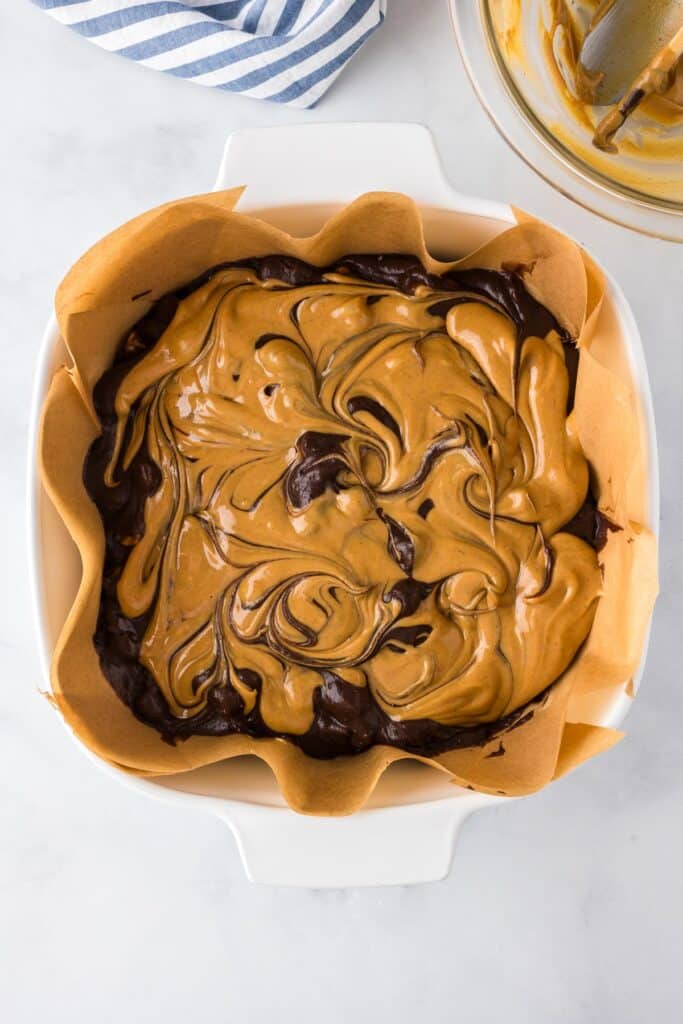 A square baking dish lined with parchment paper holds brownie batter swirled with peanut butter.