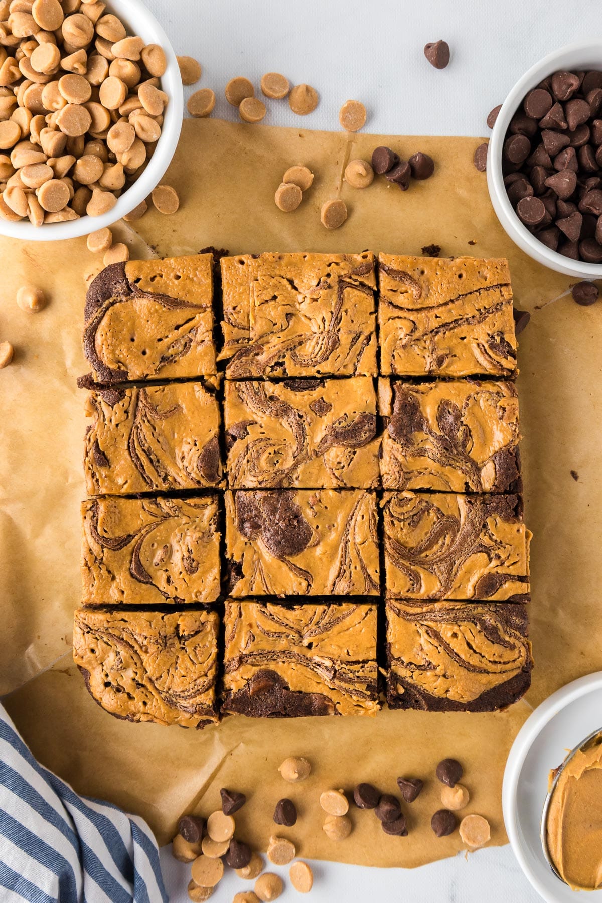 A batch of twelve peanut butter and chocolate swirl brownies sliced after baking on parchment paper, surrounded by small bowls of chocolate and peanut butter chips.