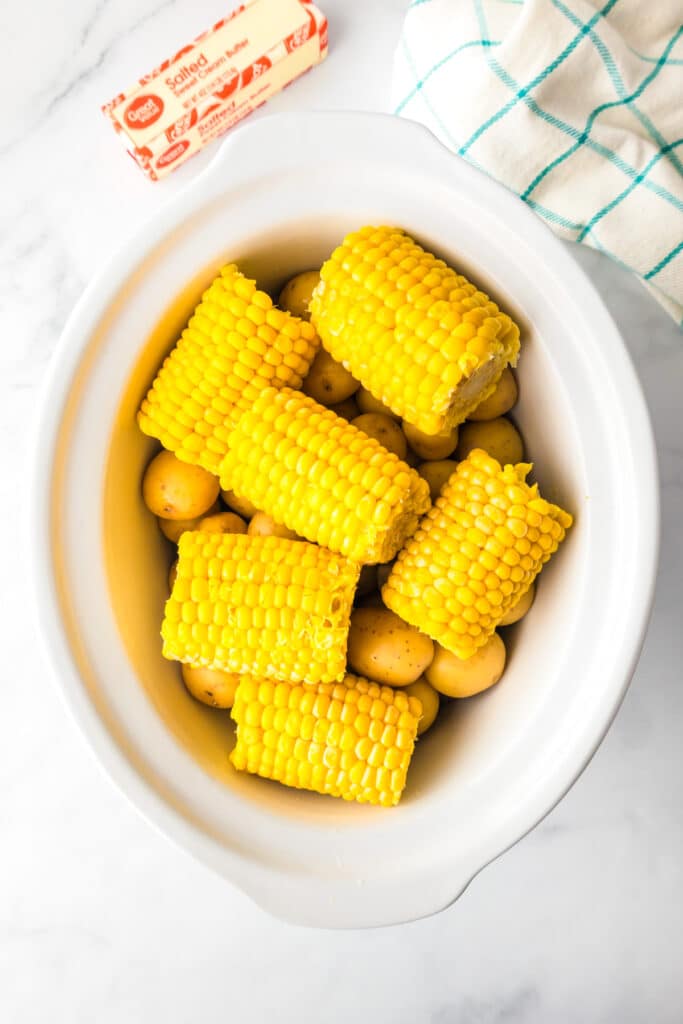 Corn pieces over small potatoes in a white slow cooker with butter on the counter nearby.