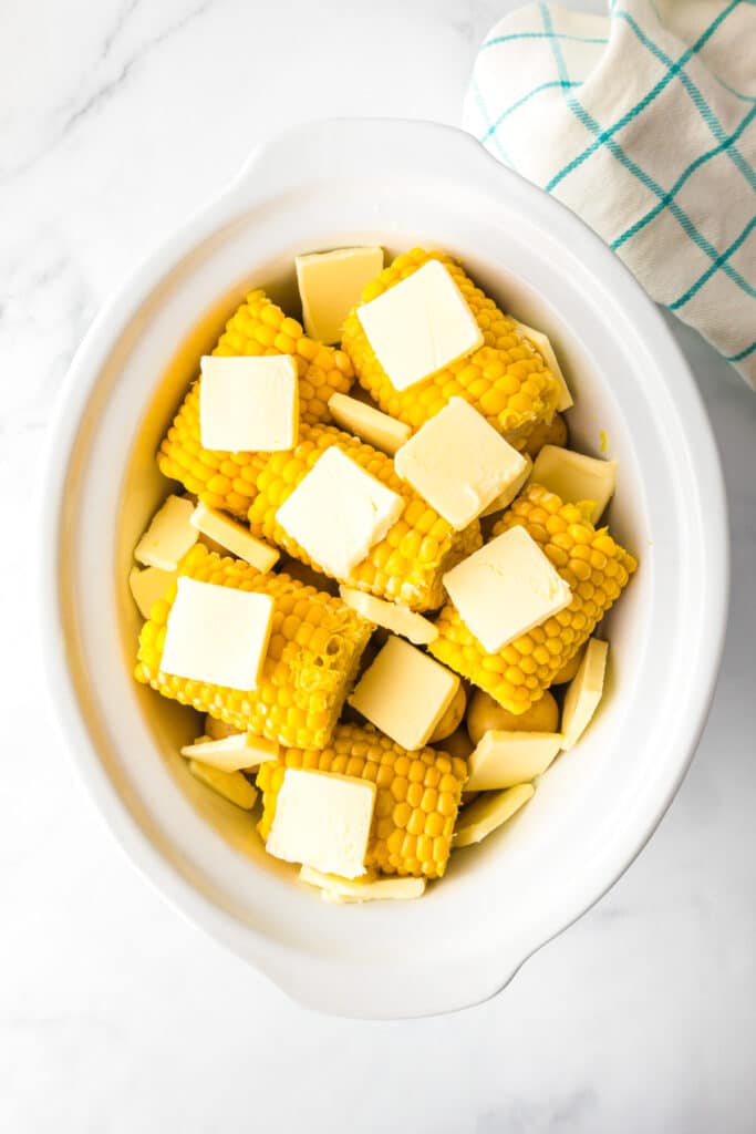 Corn on the cob pieces arranged in a white slow cooker, topped with several slices of butter over potatoes.