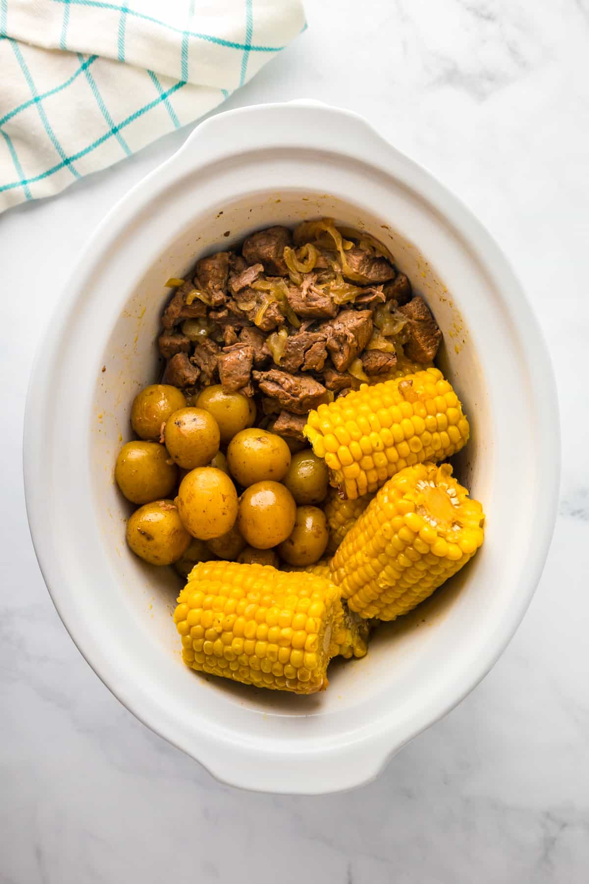 A slow cooker filled with cooked steak bites, whole baby potatoes, and corn on the cob from above.