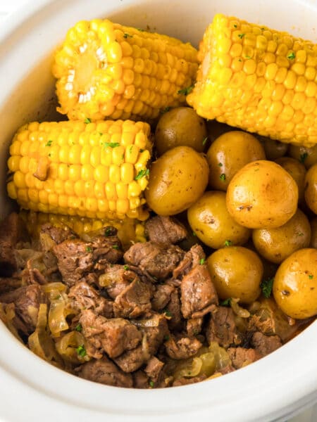 Wide view of a square view of steak pieces, potatoes and corn in a slow cooker cooked.