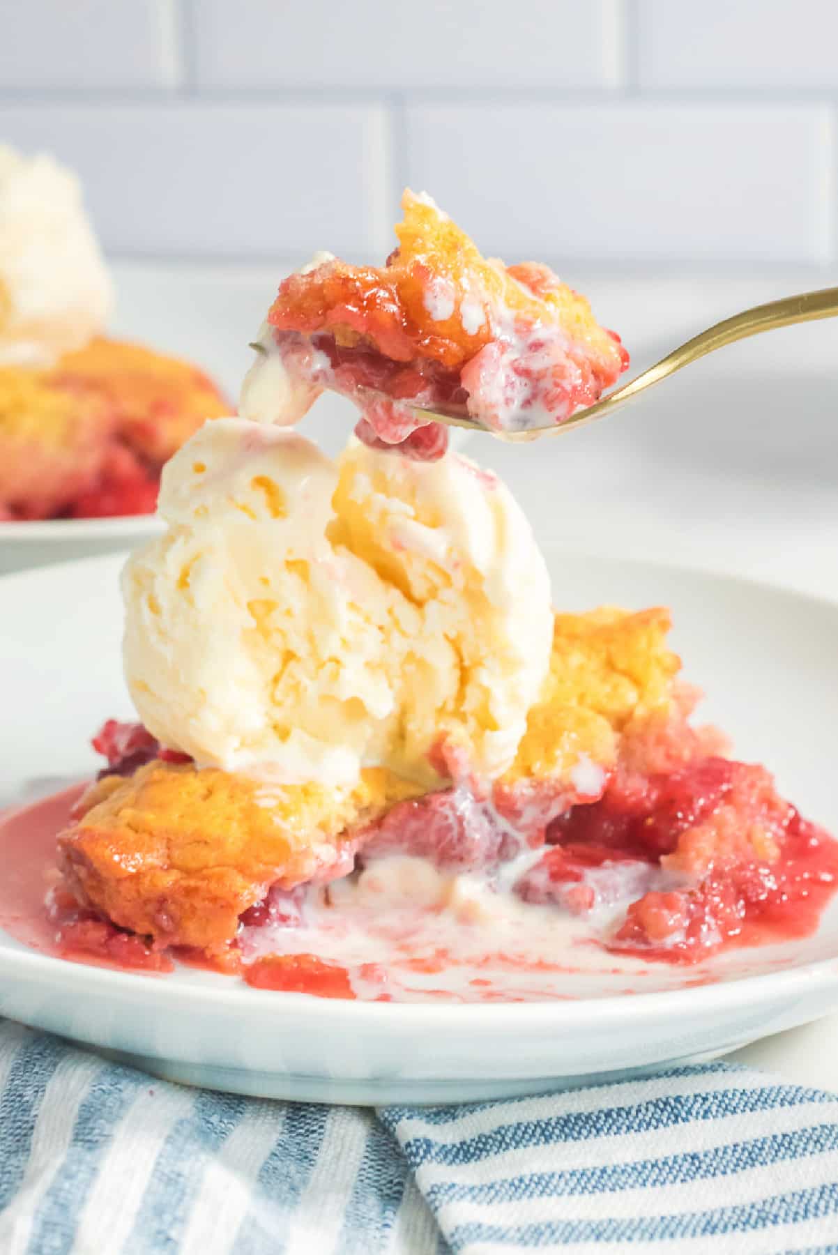 A plate with a serving of strawberry cobbler topped with a scoop of vanilla ice cream with a fork lifting a bite above the plate.