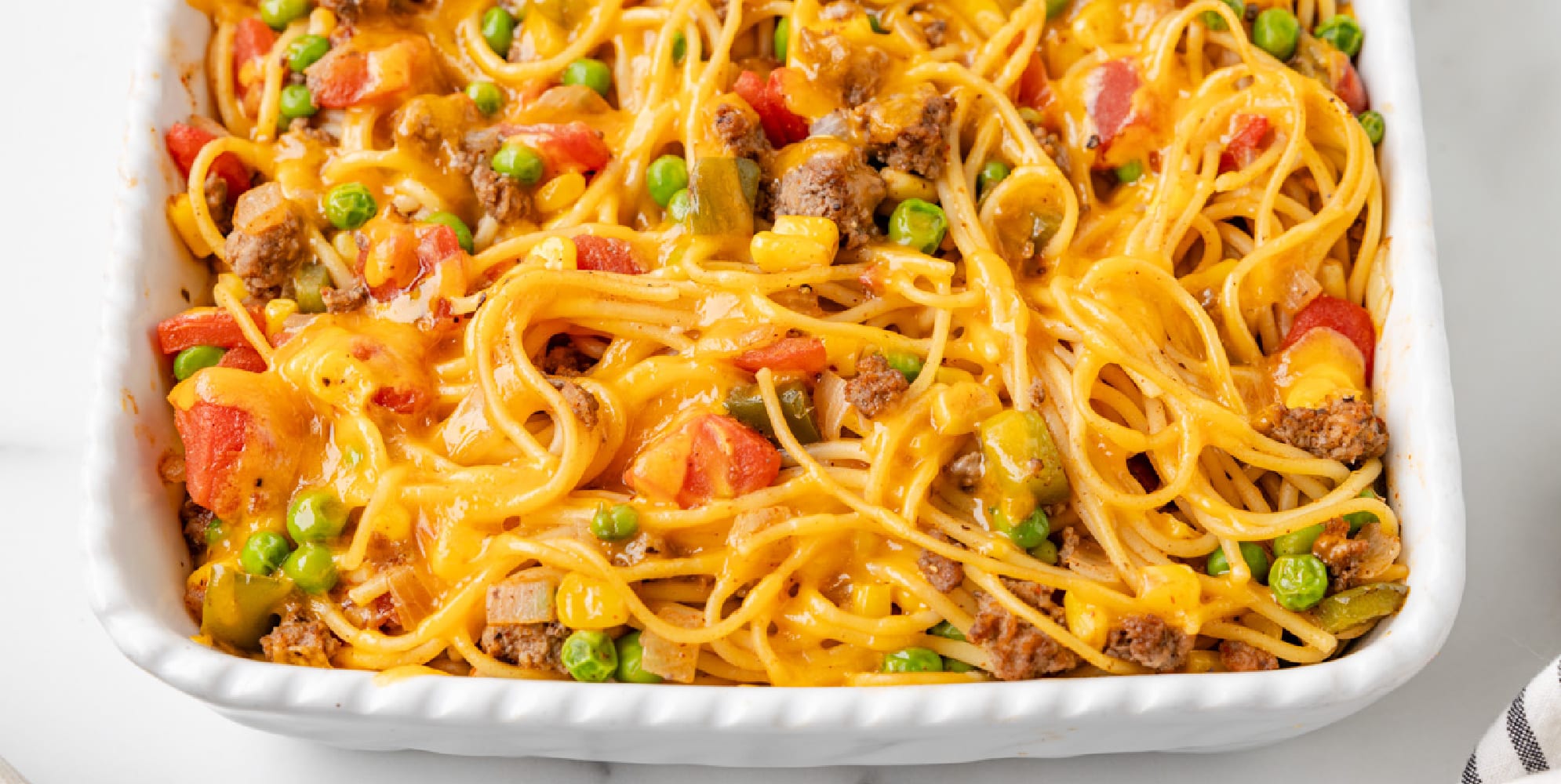 Wide view of baked church super spaghetti casserole with melted cheese, ground meat, green peas, diced tomatoes, and bell peppers in a white baking dish.