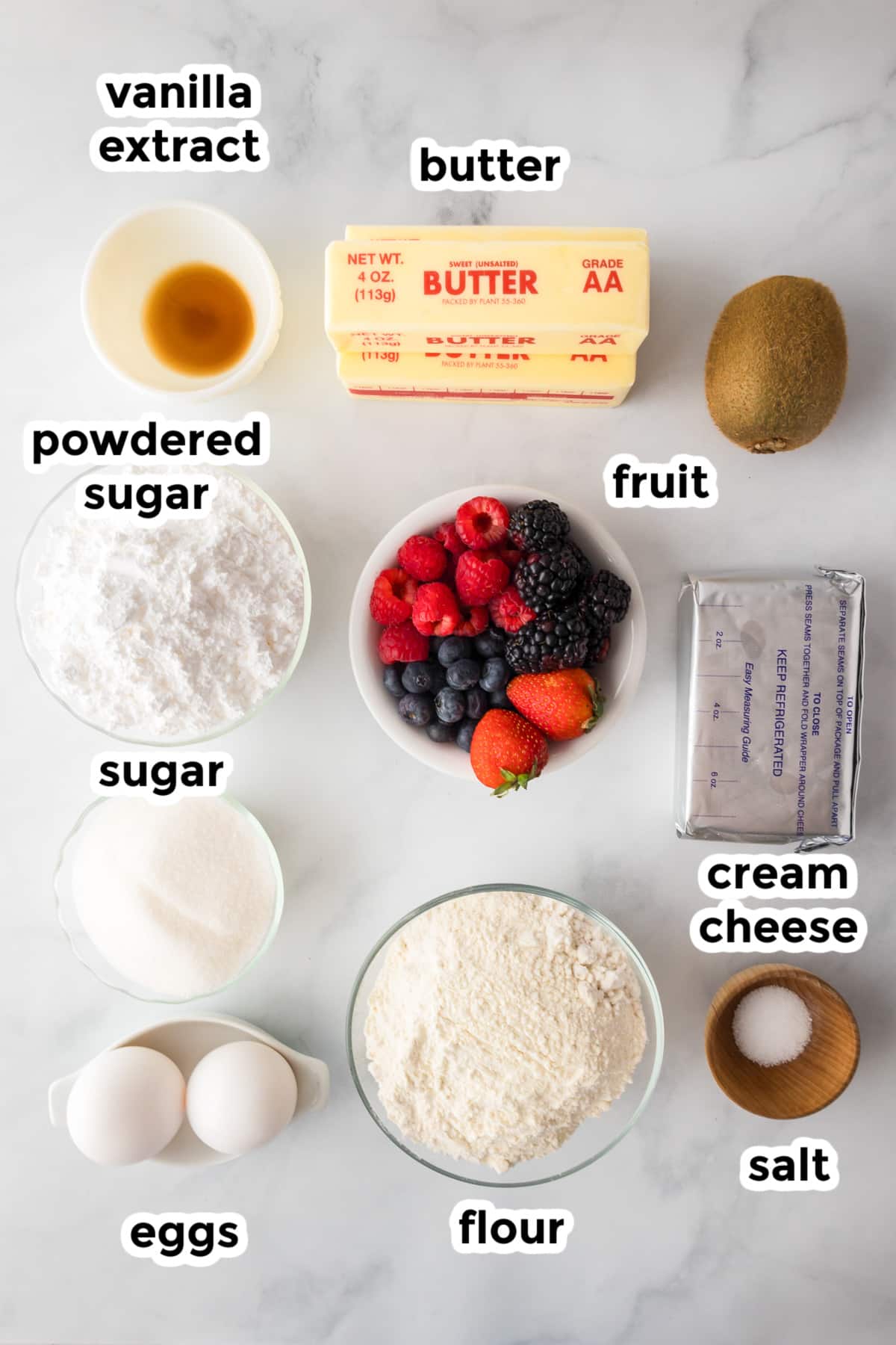 Top-down view of labeled baking ingredients: vanilla extract, butter, fruit, powdered sugar, sugar, cream cheese, flour, salt, and eggs arranged on a white surface.