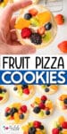 A hand holds a fruit pizza cookie topped with cream cheese frosting, kiwi, orange, raspberry, blackberry, and blueberry above a tray filled with similar fruit-topped cookies.