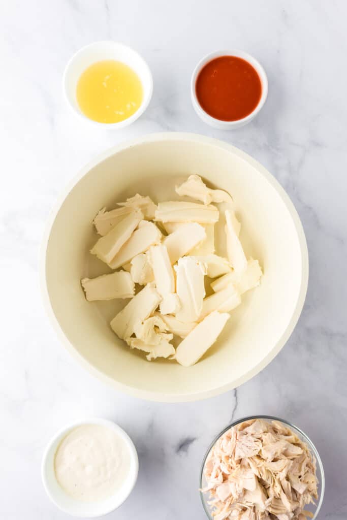 A pan of sliced cream cheese is surrounded by small bowls of chicken broth, hot sauce, shredded chicken, and ranch dressing for buffalo chicken zucchini boats.