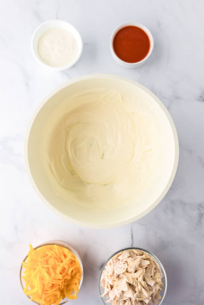 A pan full of cream cheese, surrounded by bowls of shredded cheddar cheese, cooked chicken, hot sauce, and ranch dressing for buffalo chicken zucchini boats.
