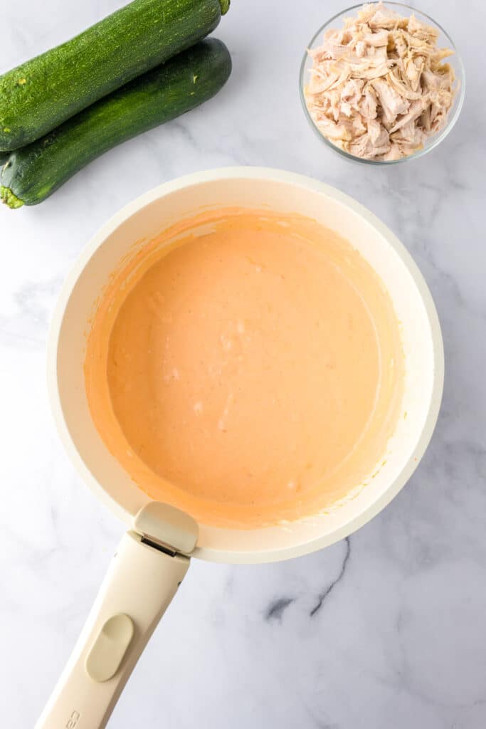 A saucepan filled with a creamy cheesy buffalo sauce to mix with chicken nearby to stuff into zucchini for buffalo chicken zucchini boats.