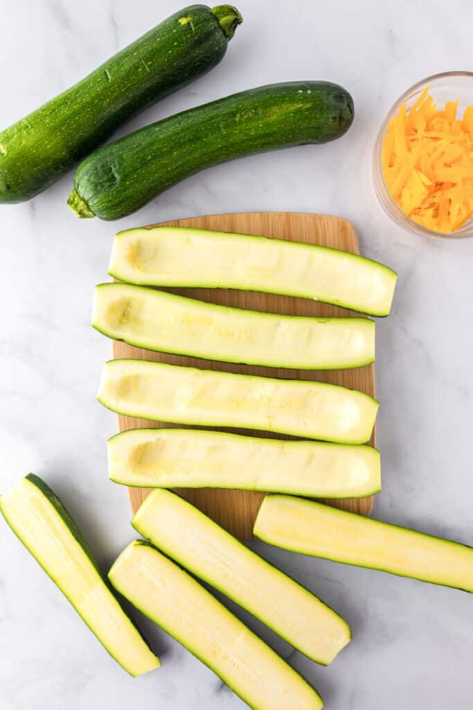 Two whole zucchinis, several halved and hollowed zucchinis on a cutting board.