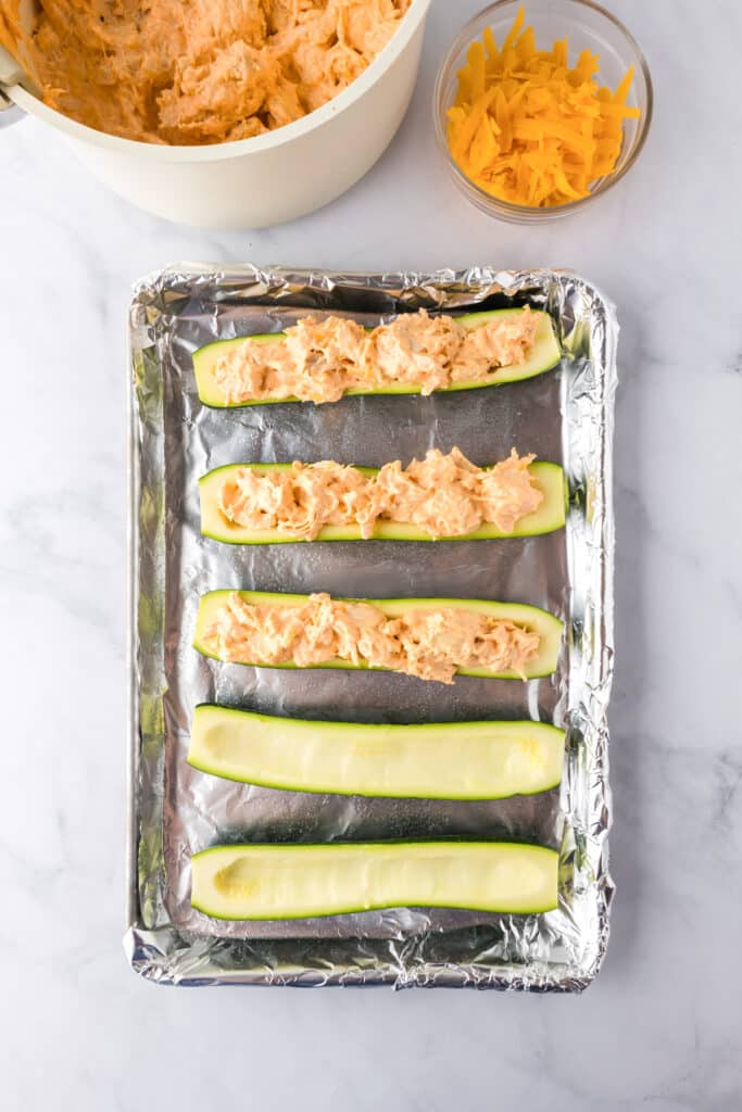 Six zucchini halves on a foil-lined baking tray, with three filled with creamy buffalo chicken.