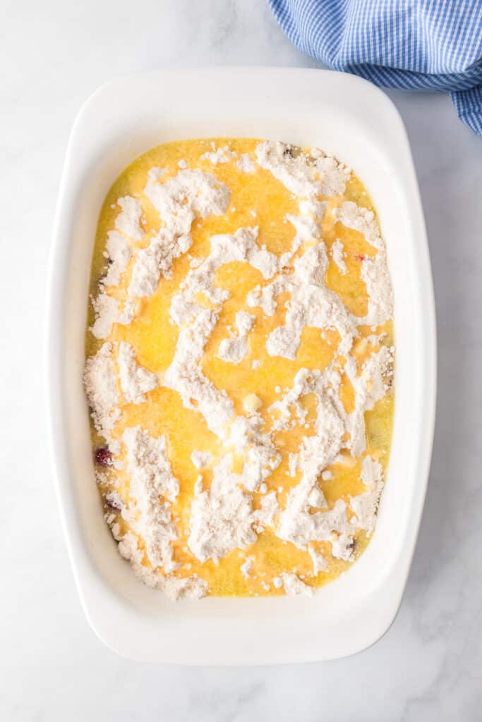 A white baking dish filled with an unbaked mixture of cake mix and melted butter over cherry pie filling on a counter.