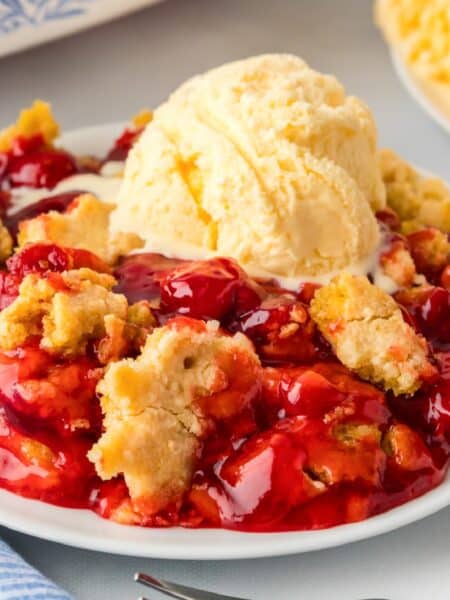 A square view of a plate of cherry cobbler topped with a scoop of melting vanilla ice cream.