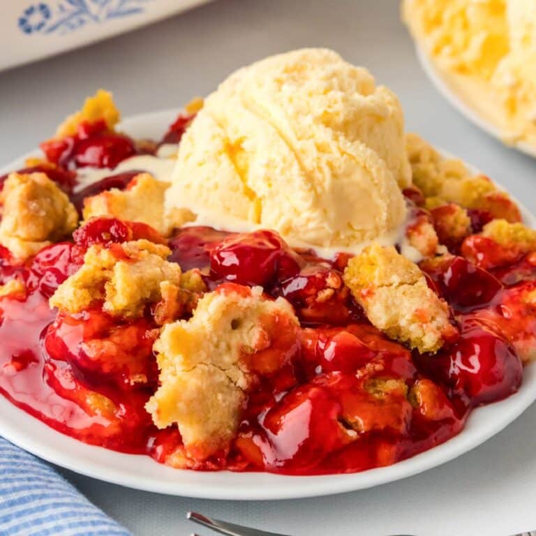 A square view of a plate of cherry cobbler topped with a scoop of melting vanilla ice cream.