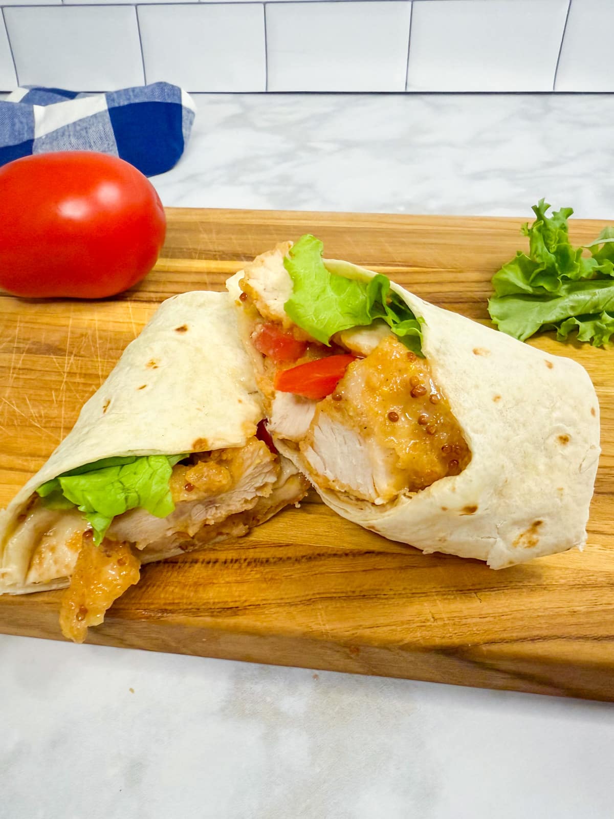 A chicken nugget wrap with lettuce, tomato, and honey mustard sauce on a wooden cutting board, with a fresh tomato and lettuce in the background.
