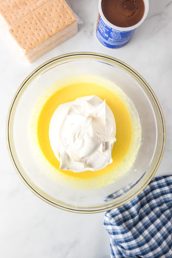 Whipped topping being folded into a mixture of instant pudding mix and milk for chocolate eclair cake in a bowl.