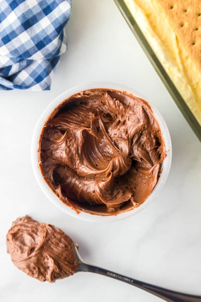A bowl of chocolate frosting sits beside a spoonful of frosting and a partially visible no bake eclair cake.