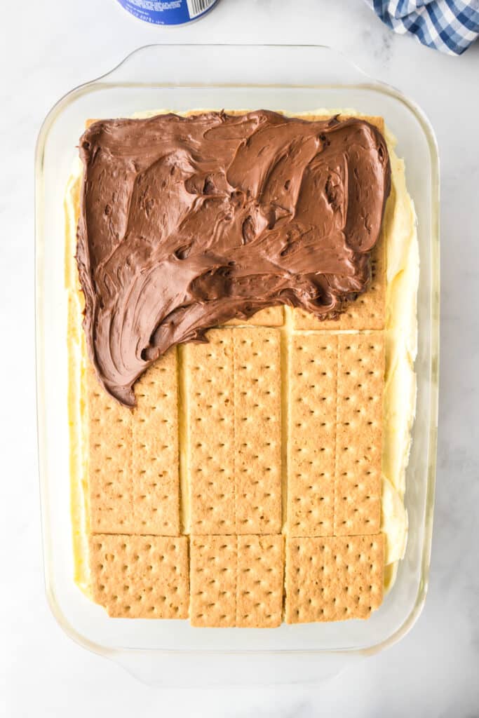 A glass dish containing a no bake chocolate eclair cake with chocolate frosting being added to the top.