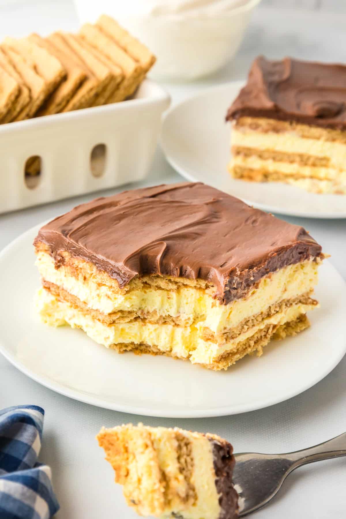 A slice of chocolate eclair cake on a plate with a bite on a fork and another slice and graham crackers in the background.