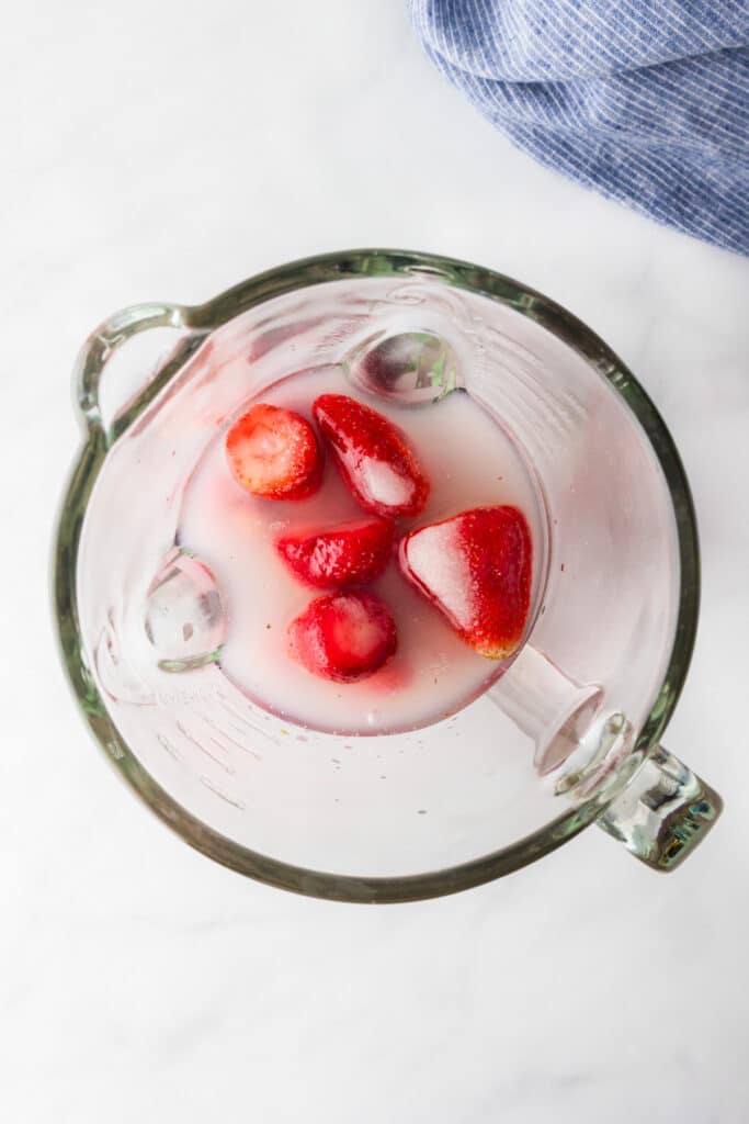 Overhead of a blender full of lemon juice, strawberries, sugar and water to blend into frozen strawberry lemonade.