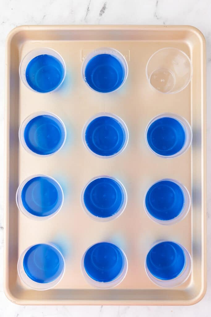 A baking sheet holds twelve plastic cups, 11 filled with blue jello from above.
