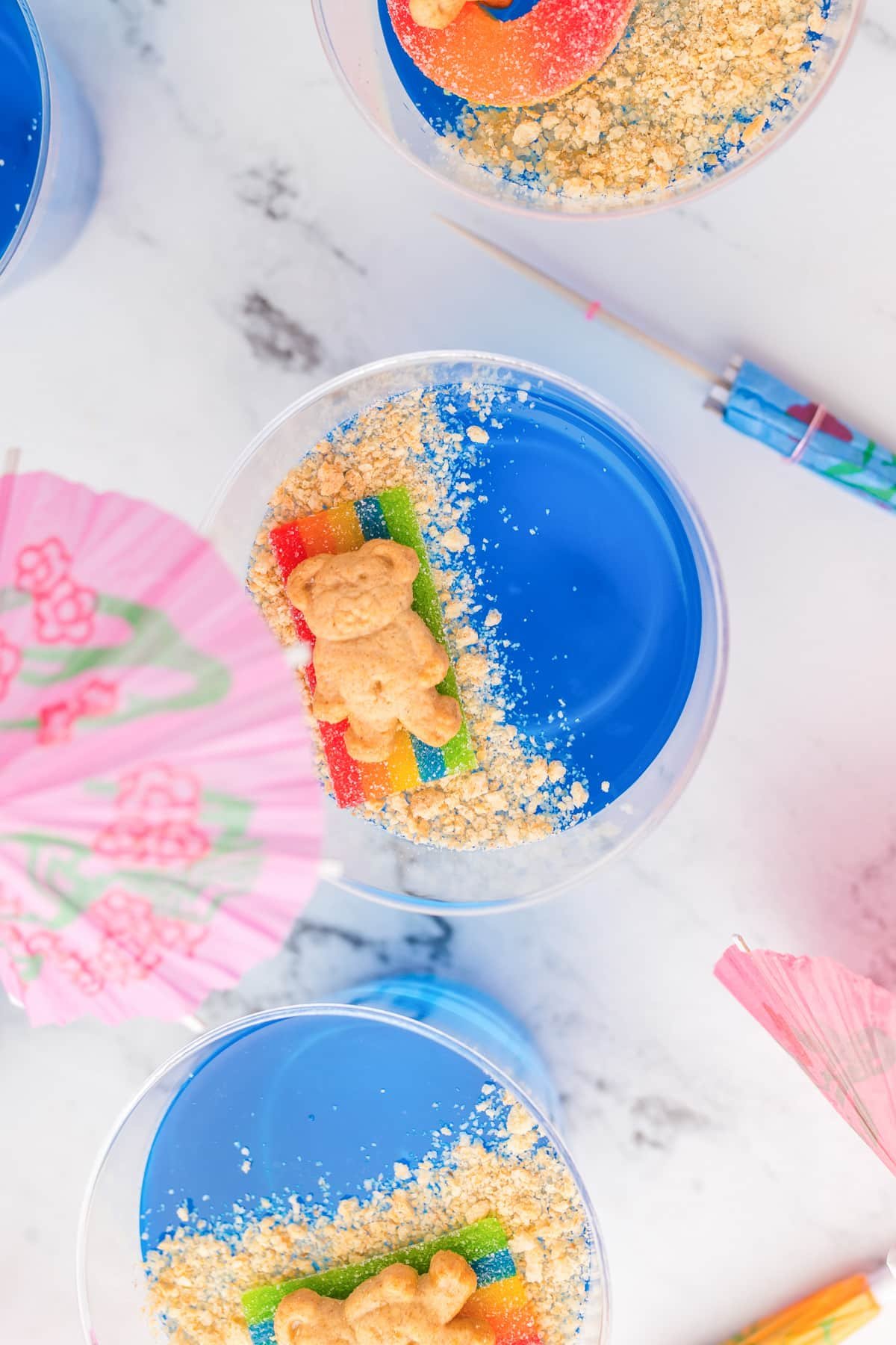 View from above of a bear cookie laying on a candy beach towel over cookie crumb sand on top of blue jello water in a cup.