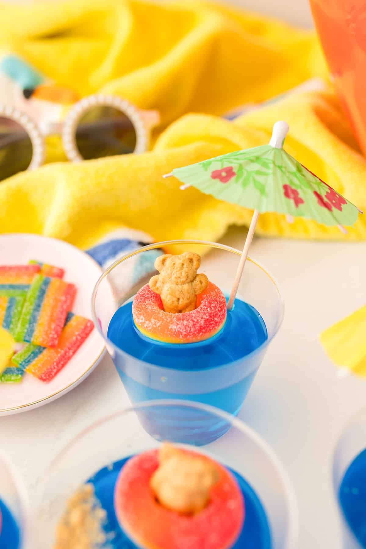A clear cup filled with blue jello decorated with a teddy graham and a candy ring to look like the bear is floating in water.