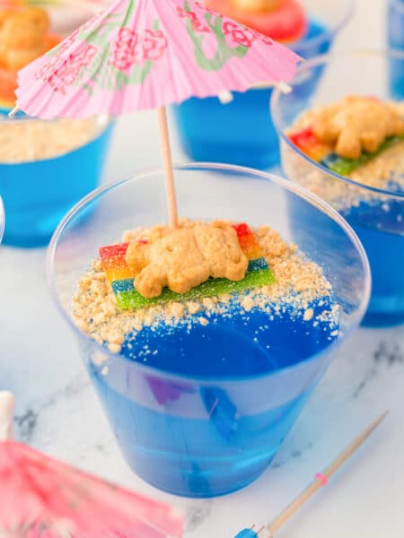Close up of a jello beach cup full of blue jello and a graham cracker bear laying on a candy towel in on the graham cracker crumb sand with more cups nearby.
