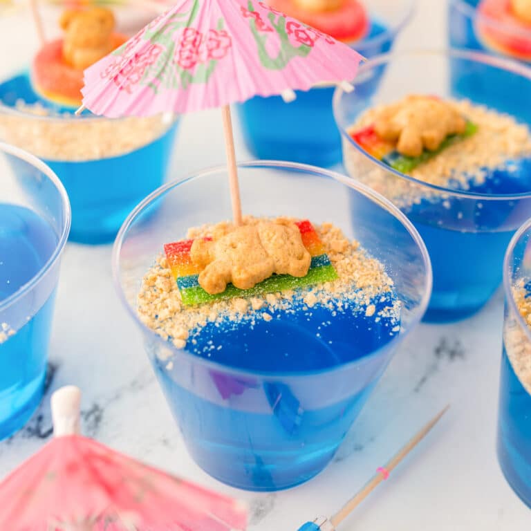 Close up of a jello beach cup full of blue jello and a graham cracker bear laying on a candy towel in on the graham cracker crumb sand with more cups nearby.