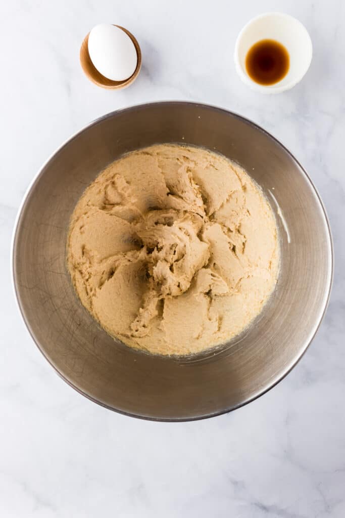 A metal mixing bowl with creamed butter and sugar for red white and blue m&m cookie dough with an egg and vanilla in bowls nearby.