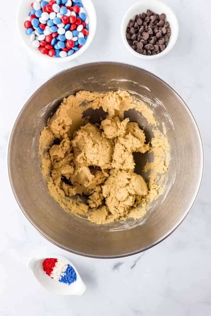 A metal bowl of cookie dough on a counter with red white and blue M&Ms, chocolate chips and sprinkles in bowls nearby.