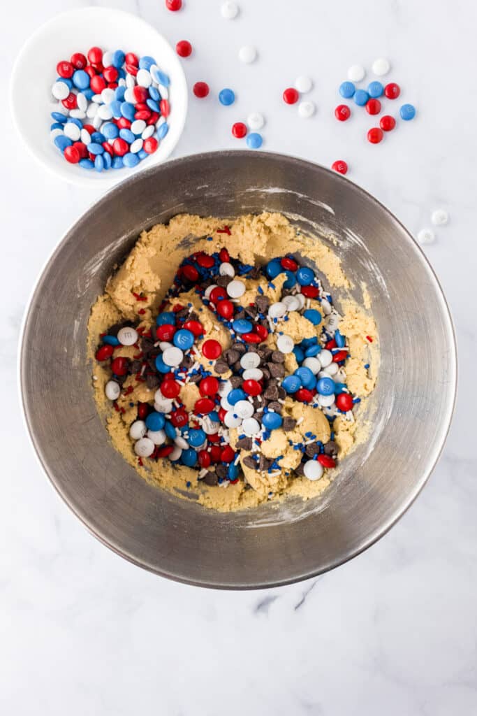 A metal mixing bowl containing cookie dough with red, white, and blue M&Ms, chocolate chips and sprinkles with a bowl of M&Ms nearby.
