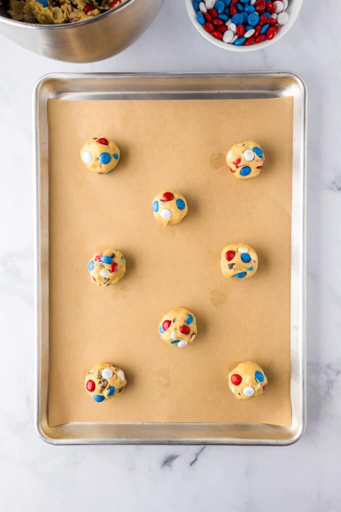 A baking sheet lined with parchment paper holds eight cookie dough balls with red, white, and blue M&Ms before baking.