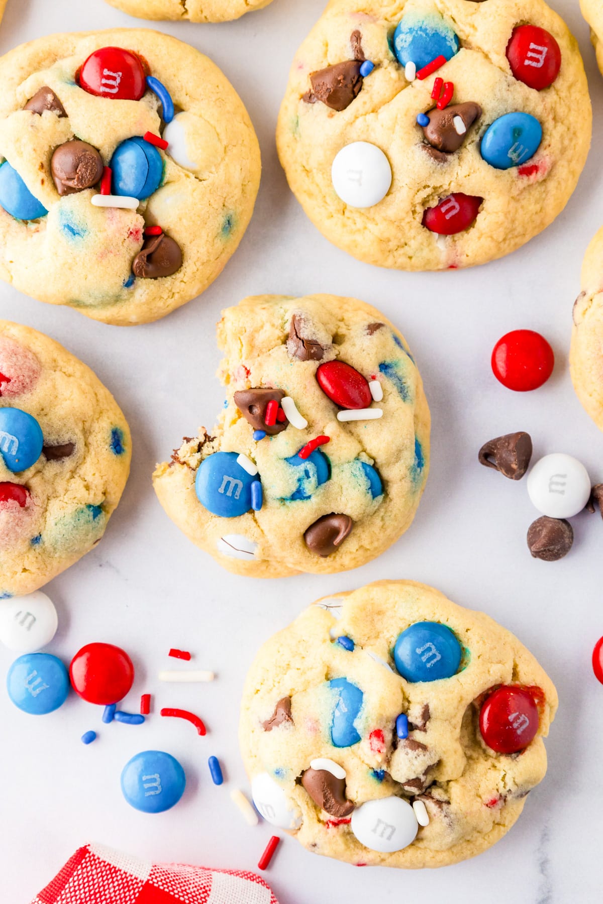 A group of cookies with red, white, and blue M&Ms, chocolate chips, and sprinkles on a counter with one cookie missing a bite.