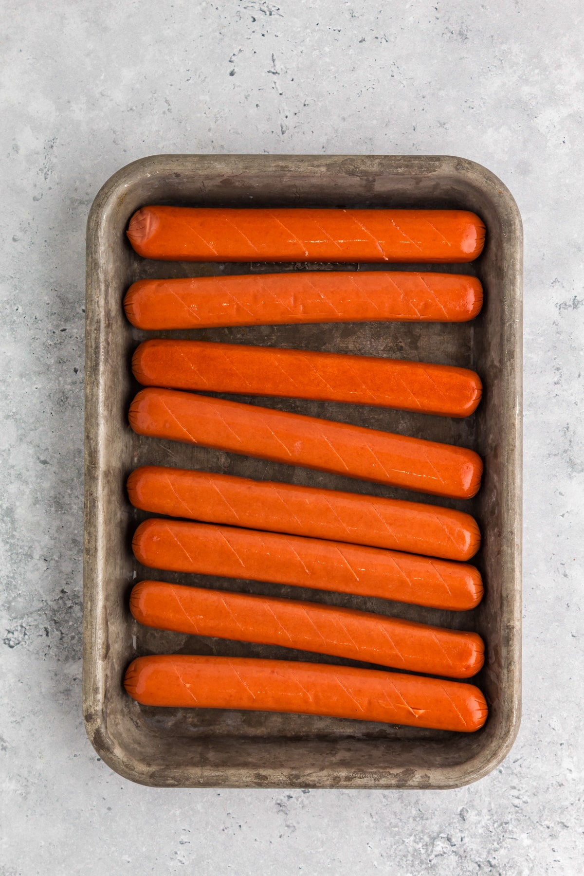 A metal baking tray with eight uncooked hot dogs that have been scoured arranged in a single layer to be wrapped in bacon.