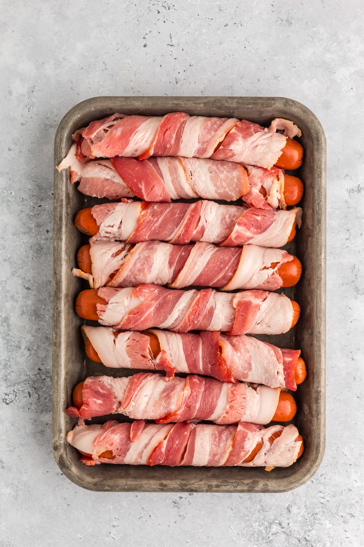 A metal tray with eight uncooked hot dogs wrapped in strips of raw bacon, arranged in a row to be put in the air fryer.